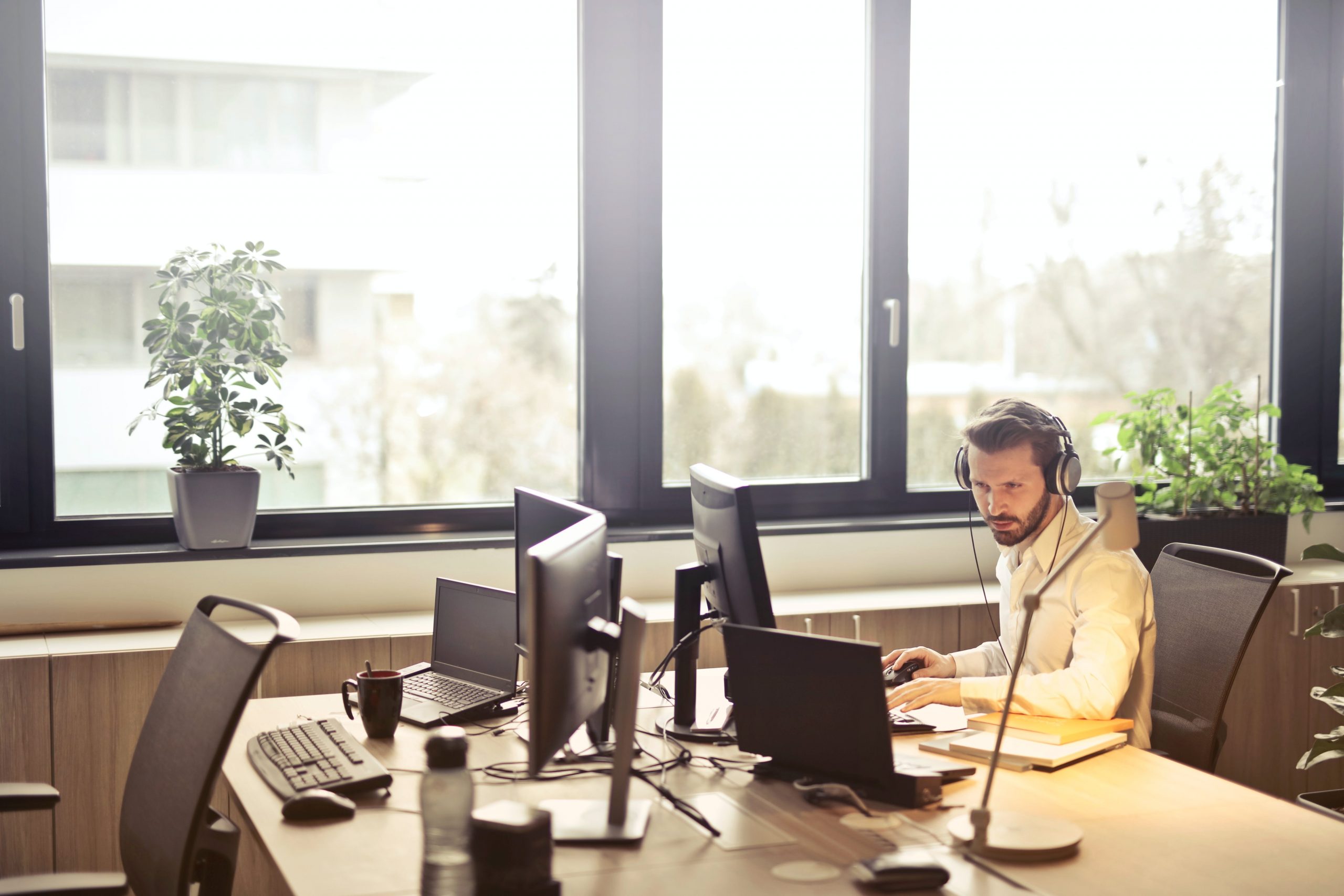 A man with a white shirt sat at a desk with a headset on surrounded by computers on a desk