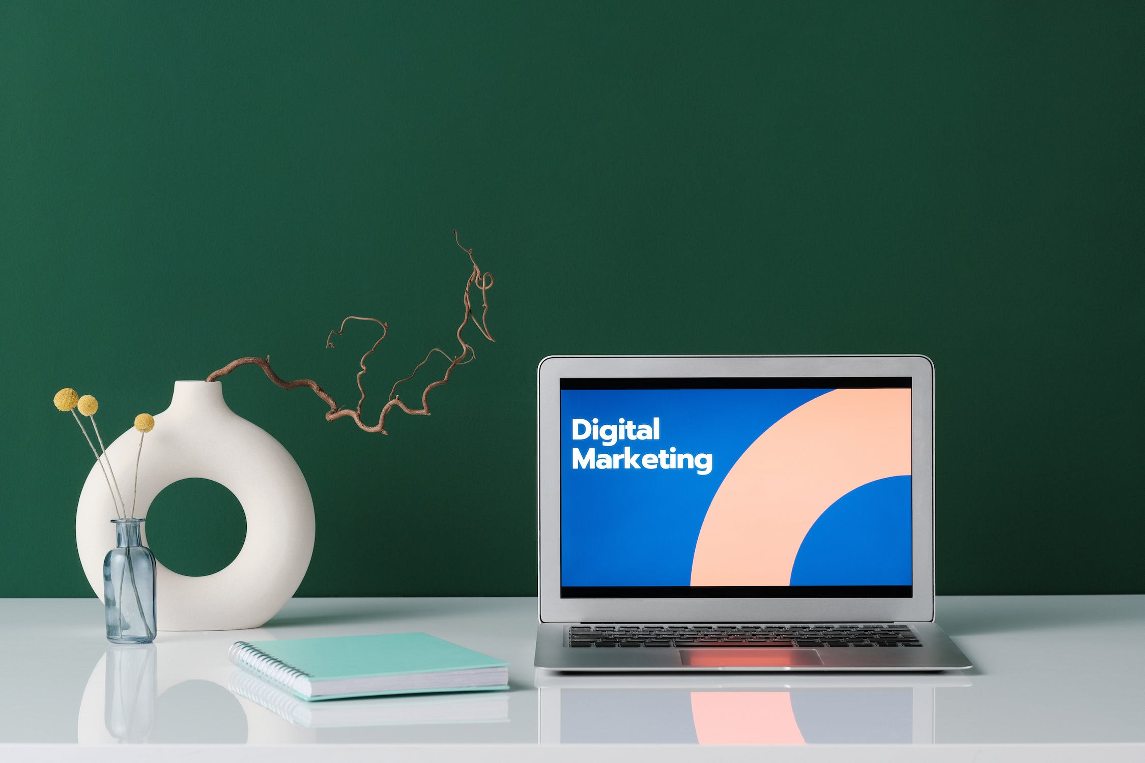 A green background with a glossy white desk with a Macbook Air laptop displaying digital marketing on the screen